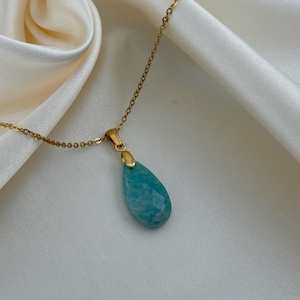 May include: A gold-colored chain necklace with a teardrop-shaped, faceted, turquoise pendant. The pendant is a light blue-green color. The necklace is displayed on a white, silky fabric.