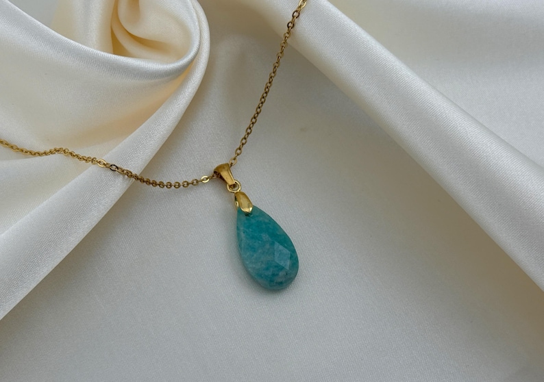 May include: A gold-colored chain necklace with a teardrop-shaped, faceted, turquoise pendant. The pendant is a light blue-green color. The necklace is displayed on a white, silky fabric.