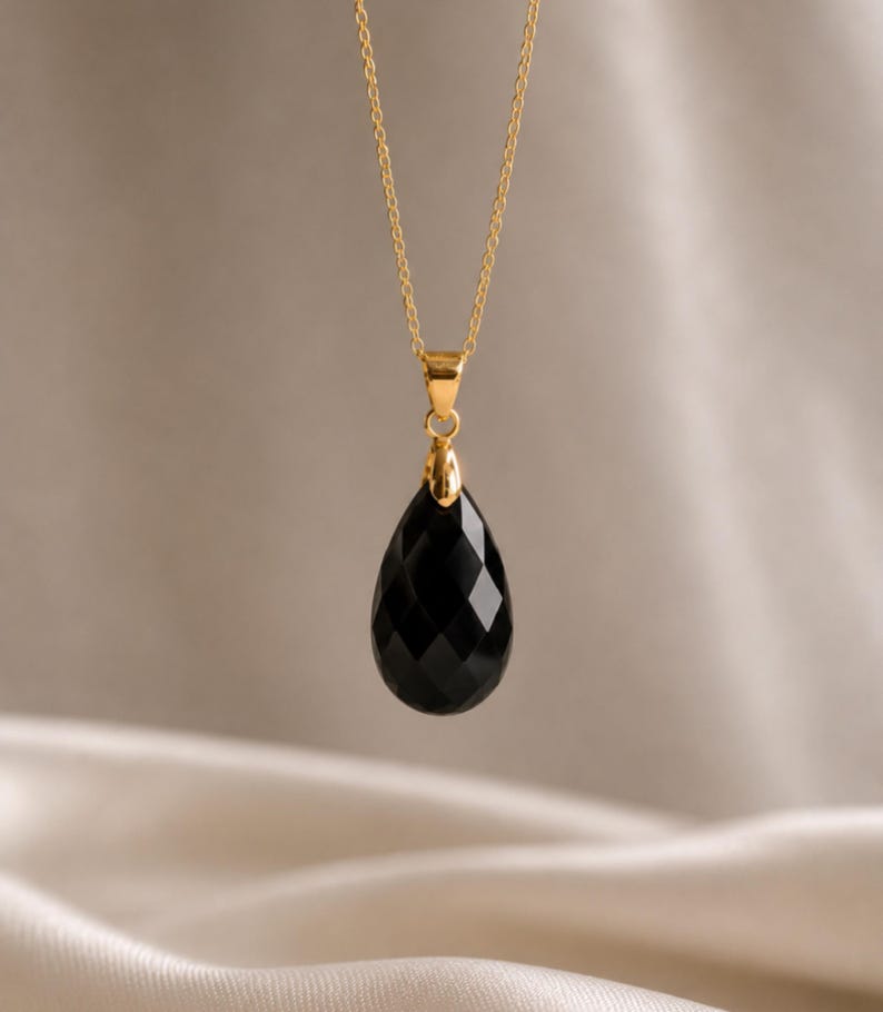 May include: A gold chain necklace with a faceted, teardrop-shaped black pendant. The pendant is suspended from a gold bail and chain, creating a simple yet elegant design. The necklace is displayed against a neutral background.