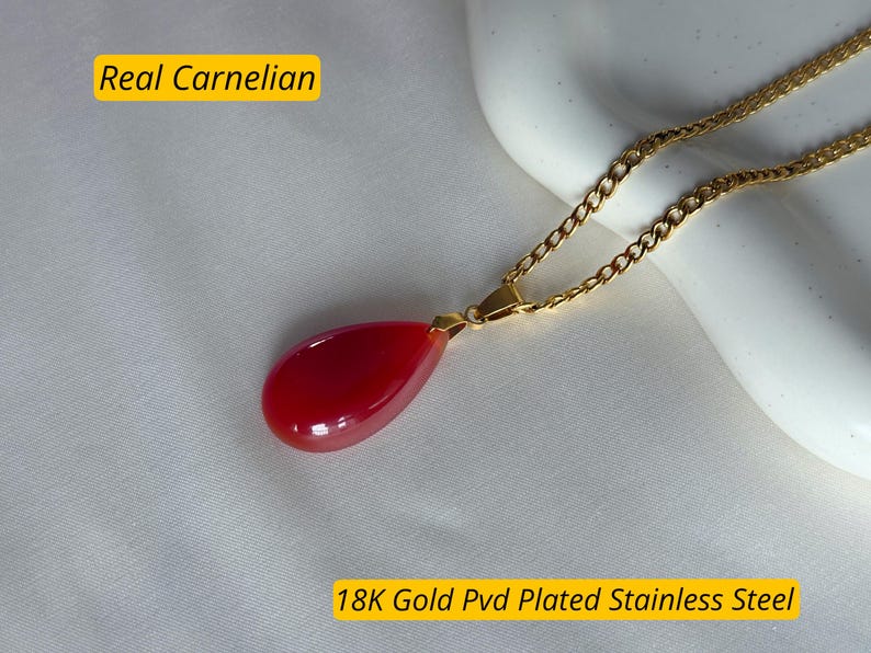 K&ouml;nnte beinhalten: Eine goldfarbene Halskette mit einem tropfenf&ouml;rmigen, leuchtend roten Karneol-Anh&auml;nger. Der Anh&auml;nger ist glatt und poliert. Der Text "Real Carnelian" und "18K Gold Pvd Plated Stainless Steel" sind im Bild.