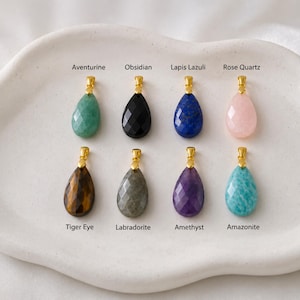 May include: Eight gemstone pendants in various colors, including green aventurine, black obsidian, blue lapis lazuli, and pink rose quartz. Each teardrop-shaped stone has a gold-colored bail. The pendants are displayed on a white, organic-shaped tray.