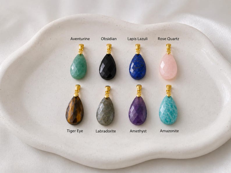 May include: Eight gemstone pendants in various colors, including green aventurine, black obsidian, blue lapis lazuli, and pink rose quartz. Each teardrop-shaped stone has a gold-colored bail. The pendants are displayed on a white, organic-shaped tray.