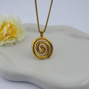 May include: A gold-colored pendant necklace featuring a spiral design. The pendant hangs from a delicate chain. A yellow and white flower rests on a white, wavy-edged tray, creating a simple, elegant composition.