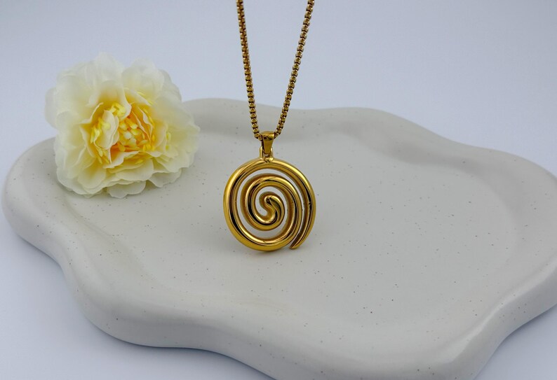 May include: A gold-colored pendant necklace featuring a spiral design. The pendant hangs from a delicate chain. A yellow and white flower rests on a white, wavy-edged tray, creating a simple, elegant composition.