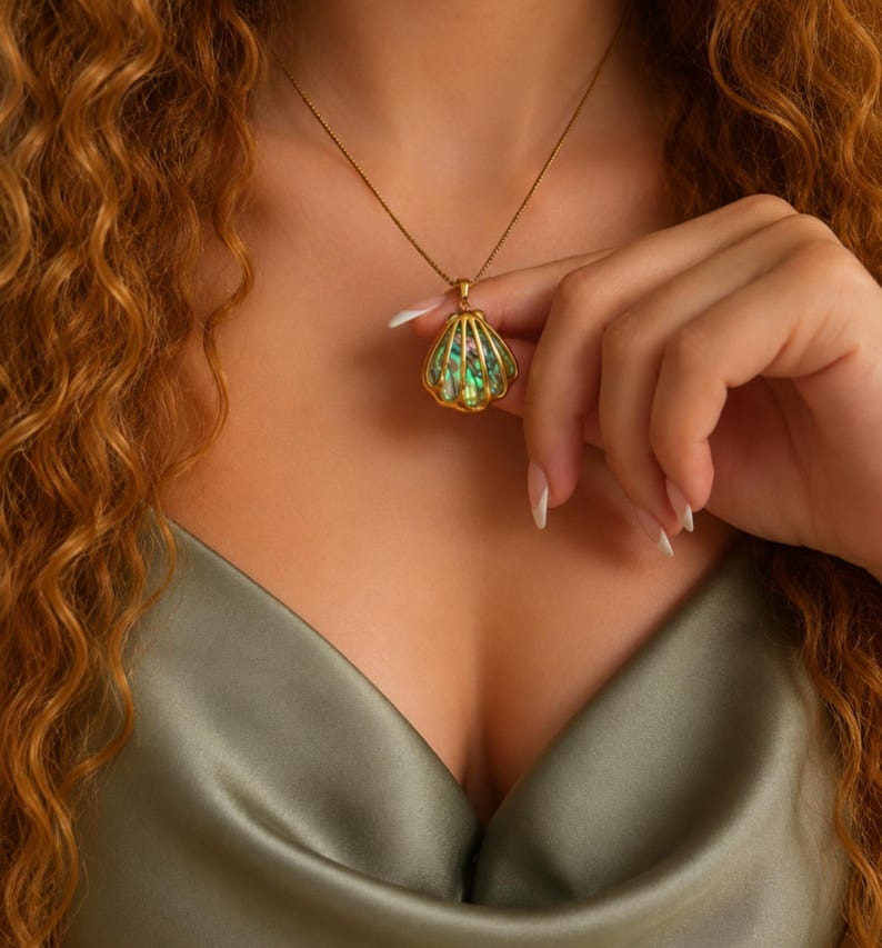 Puede incluir: Un collar dorado con un colgante de concha con acentos iridiscentes verdes y azules. El colgante cuelga de una cadena delicada. Esta joya es un collar.
