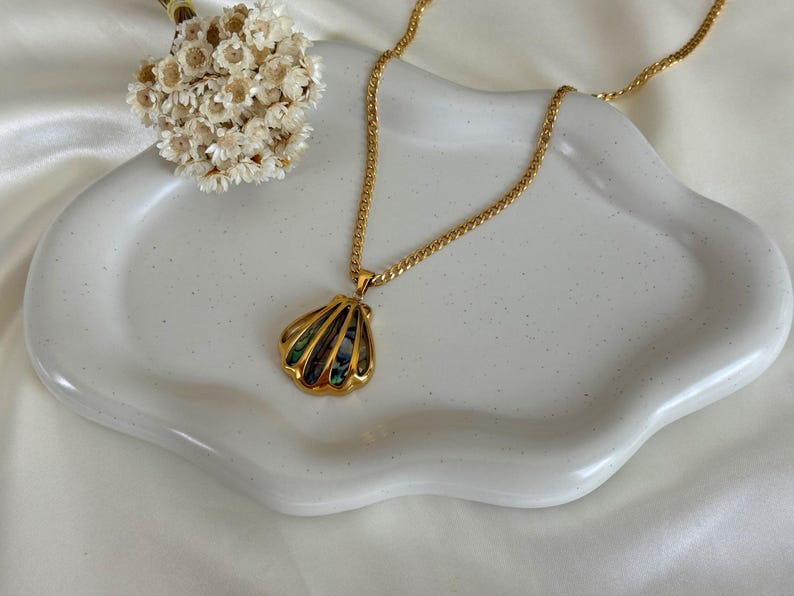 May include: Gold-toned necklace featuring a seashell pendant with iridescent accents. The necklace rests on a white, wavy-edged ceramic dish, with a small bouquet of dried flowers nearby. The chain has a textured design.