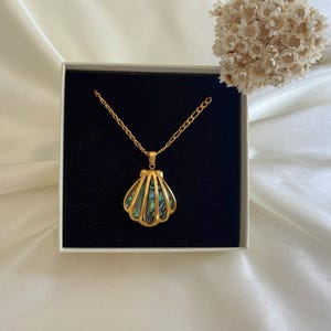 Puede incluir: Un collar con colgante de concha dorada presentado en una caja blanca con interior negro. El colgante de concha tiene acentos iridiscentes verdes y azules. El collar est&aacute; sobre una superficie de sat&eacute;n blanco, con un peque&ntilde;o ramo de flores secas en el fondo.