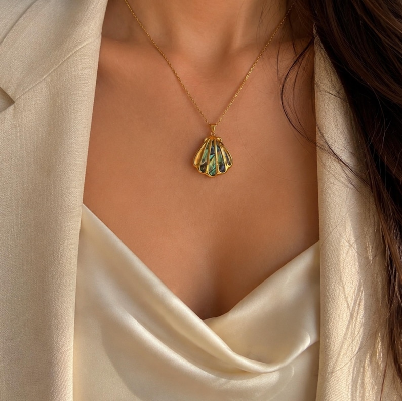 Puede incluir: Un collar con colgante de concha dorada con una cadena dorada. El colgante tiene una incrustaci&oacute;n iridiscente verde y azul. El collar se lleva sobre una blusa de sat&eacute;n color crema y una blazer beige.