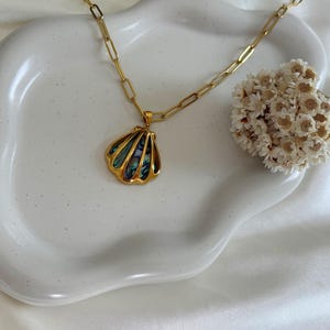 Puede incluir: Un collar dorado con un colgante de concha. El colgante presenta un contorno dorado y detalles iridiscentes azules y verdes en forma de concha. El collar est&aacute; sobre un plato de cer&aacute;mica blanco con bordes ondulados.