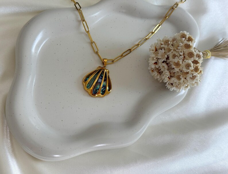 Puede incluir: Un collar dorado con un colgante de concha. El colgante presenta un contorno dorado y detalles iridiscentes azules y verdes en forma de concha. El collar est&aacute; sobre un plato de cer&aacute;mica blanco con bordes ondulados.