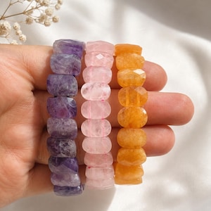 May include: Three stretch bracelets made of faceted, oval-shaped gemstone beads. The left bracelet features deep purple amethyst beads, the centre bracelet consists of pale pink rose quartz beads, and the right bracelet has vibrant orange beads.