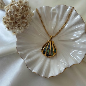 Puede incluir: Un collar dorado con un colgante de concha con incrustaciones de concha de abul&oacute;n iridiscente. El collar descansa sobre un plato blanco con borde festoneado y ribete dorado, junto a un peque&ntilde;o arreglo floral seco.