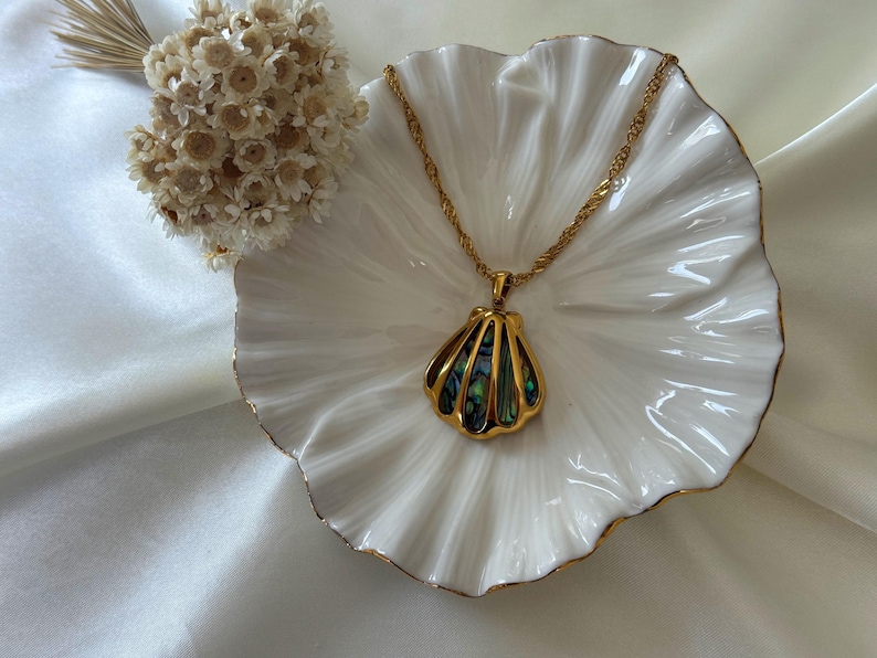 Puede incluir: Un collar dorado con un colgante de concha con incrustaciones de concha de abul&oacute;n iridiscente. El collar descansa sobre un plato blanco con borde festoneado y ribete dorado, junto a un peque&ntilde;o arreglo floral seco.