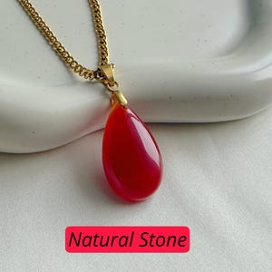 K&ouml;nnte beinhalten: Halskette mit goldfarbener Kette und einem tropfenf&ouml;rmigen, roten Anh&auml;nger aus Naturstein. Der Anh&auml;nger ist glatt und gl&auml;nzend. Der Text "Natural Stone" steht in einem roten Feld.