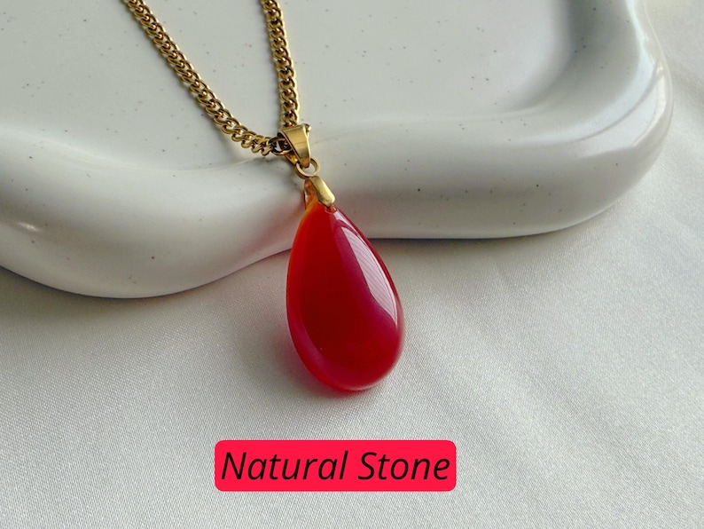 K&ouml;nnte beinhalten: Halskette mit goldfarbener Kette und einem tropfenf&ouml;rmigen, roten Anh&auml;nger aus Naturstein. Der Anh&auml;nger ist glatt und gl&auml;nzend. Der Text "Natural Stone" steht in einem roten Feld.