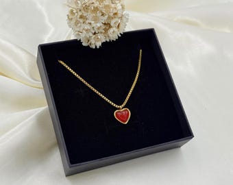 May include: A gold-toned box chain necklace featuring a small, smooth, red heart-shaped pendant with a gold-toned border. The necklace is displayed inside a square black jewelry box with a black velvet interior lining.