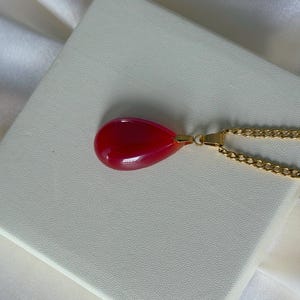 K&ouml;nnte beinhalten: Halskette mit einem tropfenf&ouml;rmigen Anh&auml;nger in leuchtendem Rot. Der Anh&auml;nger h&auml;ngt an einer goldenen Kette, die auf einer wei&szlig;en Oberfl&auml;che liegt. Ein Schmuckst&uuml;ck.