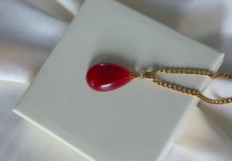 K&ouml;nnte beinhalten: Halskette mit einem tropfenf&ouml;rmigen Anh&auml;nger in leuchtendem Rot. Der Anh&auml;nger h&auml;ngt an einer goldenen Kette, die auf einer wei&szlig;en Oberfl&auml;che liegt. Ein Schmuckst&uuml;ck.