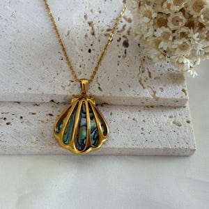 Puede incluir: Un collar dorado con un colgante de concha. El colgante presenta un marco dorado con un dise&ntilde;o iridiscente y multicolor de concha de abul&oacute;n. El collar se exhibe sobre una superficie clara y texturizada, con flores secas en el fondo.