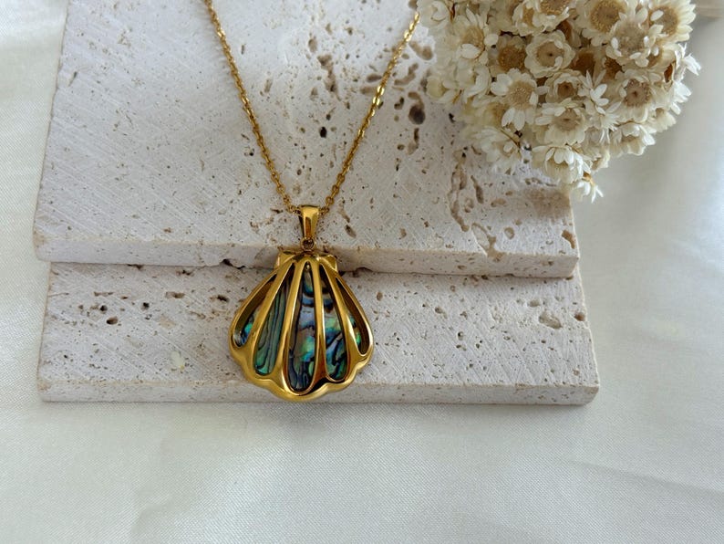 Puede incluir: Un collar dorado con un colgante de concha. El colgante presenta un marco dorado con un dise&ntilde;o iridiscente y multicolor de concha de abul&oacute;n. El collar se exhibe sobre una superficie clara y texturizada, con flores secas en el fondo.