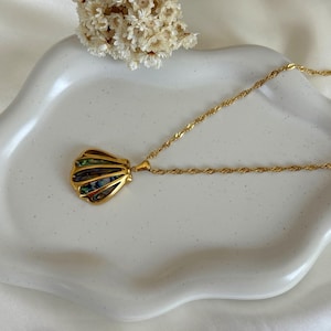 Puede incluir: Collar con colgante de concha dorada. El colgante presenta un dise&ntilde;o detallado de concha con acentos iridiscentes. El collar tiene una cadena delicada. La joya se exhibe en un plato blanco con bordes ondulados.