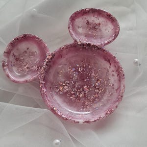 Pu&ograve; includere: Un portagioie in resina rosa a forma di orecchie di Topolino. Il piatto &egrave; riempito di glitter e scaglie iridescenti. Il piatto &egrave; su una superficie bianca, strutturata con perle sparse.