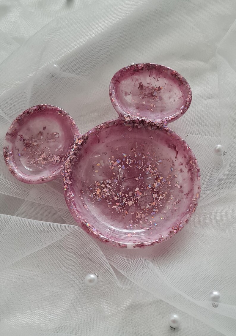 Pu&ograve; includere: Un portagioie in resina rosa a forma di orecchie di Topolino. Il piatto &egrave; riempito di glitter e scaglie iridescenti. Il piatto &egrave; su una superficie bianca, strutturata con perle sparse.