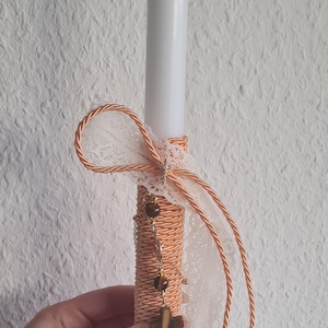 May include: A white candle decorated with orange twine, lace, and a cross pendant. The candle is adorned with a decorative bow and a chain with brown beads. The candle is held against a textured white wall.