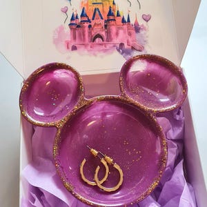 Pu&ograve; includere: Un portagioie in resina viola a forma di orecchie di Topolino, con dettagli glitterati dorati. Il piatto contiene orecchini a cerchio dorati e si trova in una scatola con un'illustrazione del castello Disney.