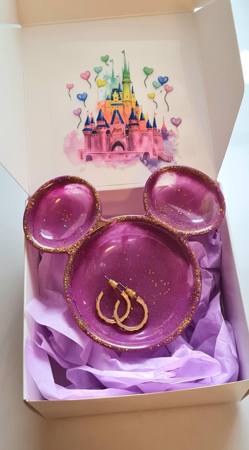 Pu&ograve; includere: Un portagioie in resina viola a forma di orecchie di Topolino, con dettagli glitterati dorati. Il piatto contiene orecchini a cerchio dorati e si trova in una scatola con un'illustrazione del castello Disney.