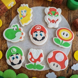 Könnte beinhalten: Eine Sammlung von acht verzierten Keksen mit Charakteren und Gegenständen aus der Super Mario-Reihe, darunter Mario, Luigi, Prinzessin Peach, Bowser, Yoshi, eine Piranha-Pflanze, eine Feuerblume und ein roter Pilz auf einer runden Unterlage.