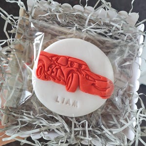 Peut inclure: Un biscuit blanc avec un motif de voiture rouge et le nom "LIAM" au centre. Le biscuit est emballé dans un sac en plastique transparent, entouré de papier déchiqueté argenté, dans une boîte blanche.