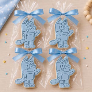 Könnte beinhalten: Vier hellblaue Kekse in Form einer Figur in transparenten Zellophanbeuteln, gebunden mit hellblauen Satinbändern. Die Kekse sind wie ein Cartoon-Hund geformt. Auf einer hellen Holzoberfläche.