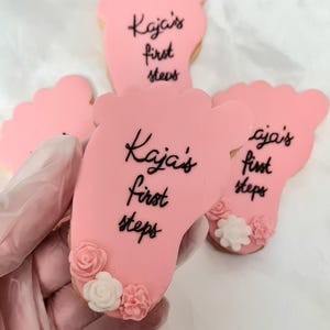 Puede incluir: Galletas rosas con forma de pie con el texto "Kaja's first steps" en escritura negra. Las galletas están decoradas con pequeños detalles florales rosas y blancos. Una mano enguantada sostiene una de las galletas.