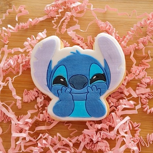 Könnte beinhalten: Ein dekorierter Zuckerkeks in Form der Disney-Figur Stitch. Der Keks hat blaue, weiße und schwarze Glasur mit Stitchs charakteristischen großen Ohren und Gesichtszügen. Der Keks liegt auf einer Holzoberfläche mit rosa Papierstreuseln.