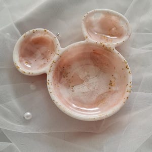 Pu&ograve; includere: Un piatto decorativo a tre sezioni a forma di testa di topo. Il piatto &egrave; bianco con un interno marmorizzato rosa e bianco e pagliuzze dorate. Il piatto &egrave; probabilmente realizzato in resina o ceramica e si trova su un tessuto bianco trasparente.