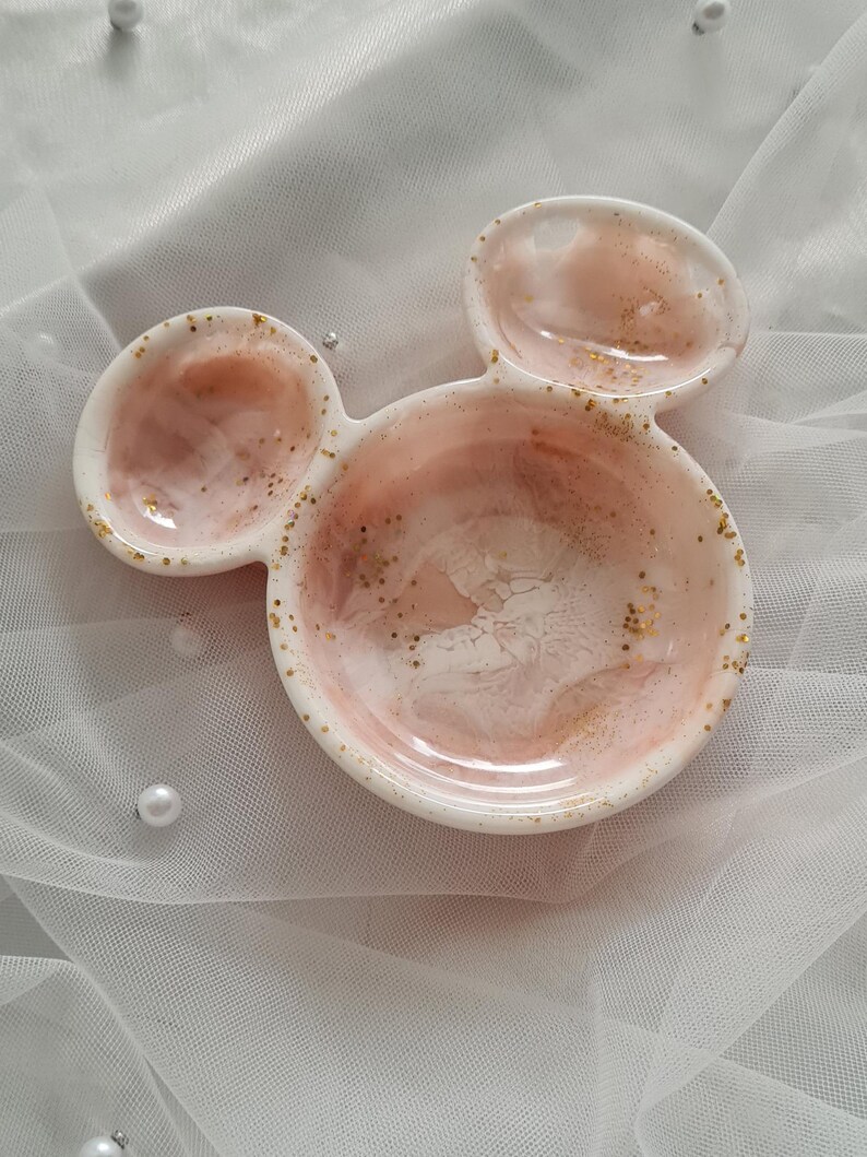 Pu&ograve; includere: Un piatto decorativo a tre sezioni a forma di testa di topo. Il piatto &egrave; bianco con un interno marmorizzato rosa e bianco e pagliuzze dorate. Il piatto &egrave; probabilmente realizzato in resina o ceramica e si trova su un tessuto bianco trasparente.