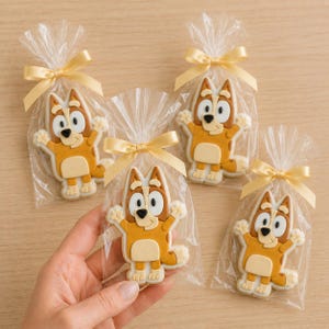 May include: Four individually wrapped sugar cookies shaped like the cartoon character Bluey, colored in shades of tan and cream with black details. Each cookie is sealed in a clear plastic bag tied with a light yellow satin ribbon bow.