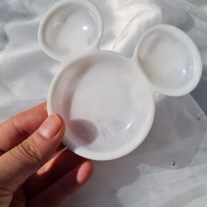 Pu&ograve; includere: Un piatto bianco a forma di Topolino. Il piatto ha una finitura lucida ed &egrave; tenuto in mano. Il piatto &egrave; su una superficie bianca strutturata con perle sparse.