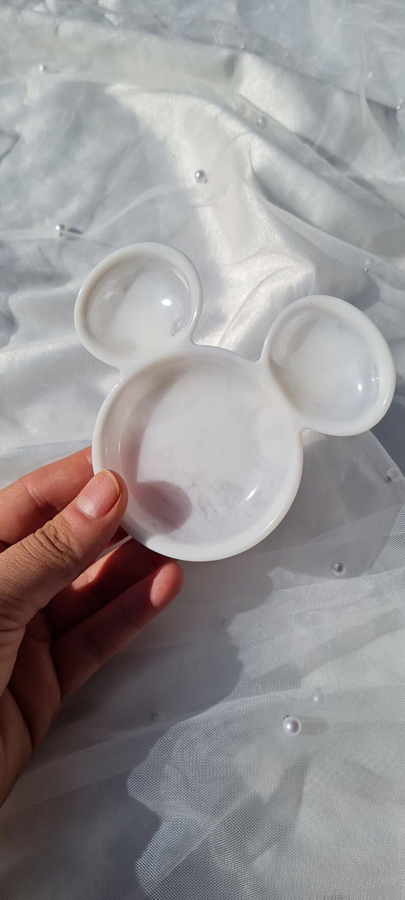 Pu&ograve; includere: Un piatto bianco a forma di Topolino. Il piatto ha una finitura lucida ed &egrave; tenuto in mano. Il piatto &egrave; su una superficie bianca strutturata con perle sparse.