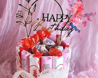 May include: A festive birthday cake made of pink and white chocolate bars, decorated with a gold "Happy Birthday" topper and a lace ribbon. The cake is filled with candies and wrapped in a pink fabric.