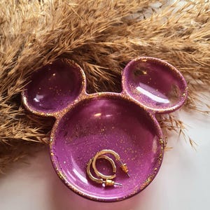 Pu&ograve; includere: Un vassoio viola a forma di testa di Topolino, con macchie e bordi dorati. Il vassoio contiene un paio di orecchini a cerchio dorati. Erbe secche sullo sfondo.