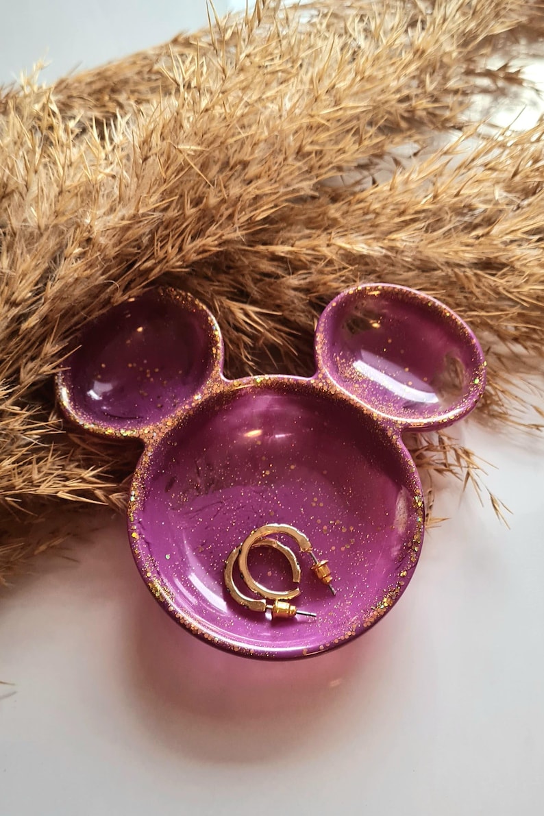 Pu&ograve; includere: Un vassoio viola a forma di testa di Topolino, con macchie e bordi dorati. Il vassoio contiene un paio di orecchini a cerchio dorati. Erbe secche sullo sfondo.