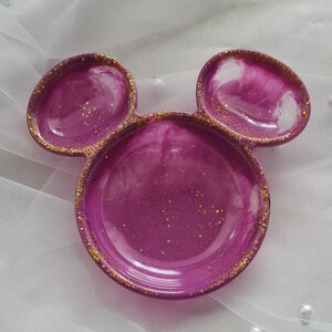 Pu&ograve; includere: Un portagioie decorativo a forma di testa di Topolino. Il piatto &egrave; di un vibrante colore magenta con accenti di glitter dorati sui bordi e all'interno della resina. Su uno sfondo bianco strutturato.