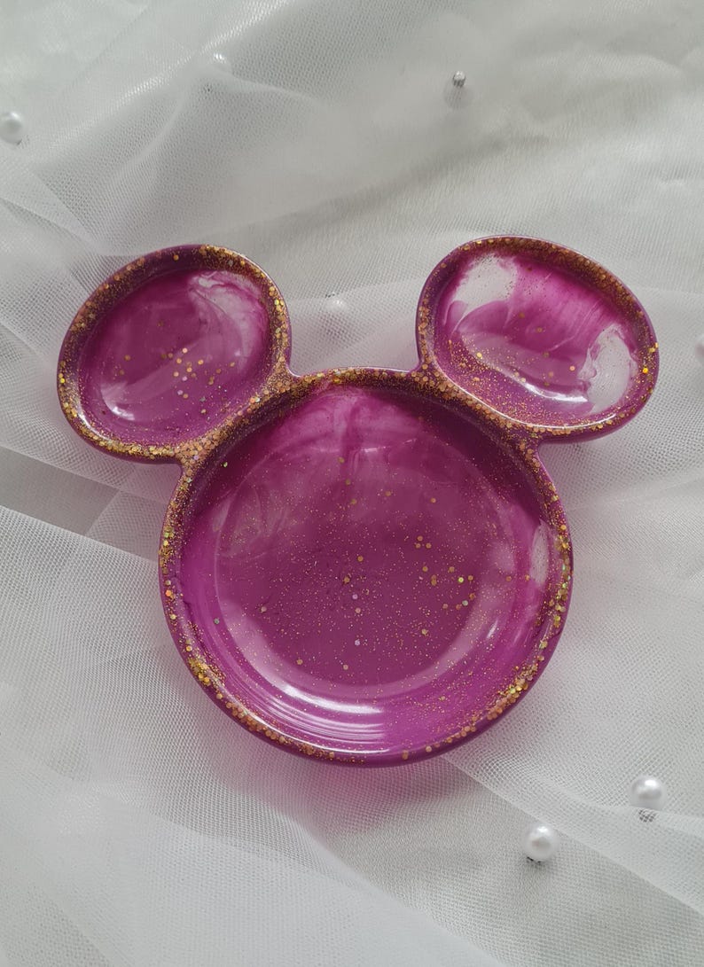 Pu&ograve; includere: Un portagioie decorativo a forma di testa di Topolino. Il piatto &egrave; di un vibrante colore magenta con accenti di glitter dorati sui bordi e all'interno della resina. Su uno sfondo bianco strutturato.