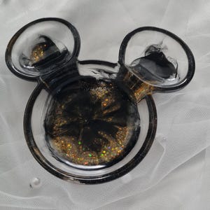 Pu&ograve; includere: Un portagioie a forma di Mickey Mouse in resina. Il piatto &egrave; nero e trasparente con glitter dorati e vortici neri. Ha due orecchie rotonde e una base circolare.