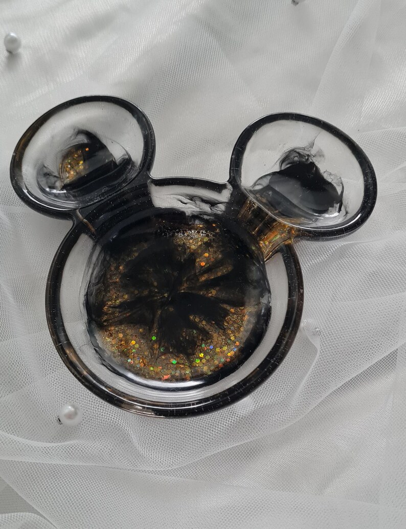 Pu&ograve; includere: Un portagioie a forma di Mickey Mouse in resina. Il piatto &egrave; nero e trasparente con glitter dorati e vortici neri. Ha due orecchie rotonde e una base circolare.