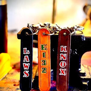 May include: Three leather keychains in black, orange, and brown. The black keychain has the word "LAWS" in white and red lettering. The orange keychain has the numbers "E132" in green. The brown keychain has the word "KNOX" in white and red.
