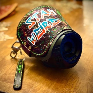 May include: A black and blue device with a leather cover featuring the words "STAY WEIRD" in colorful graffiti-style lettering. A small black leather keychain with green text hangs from the device.