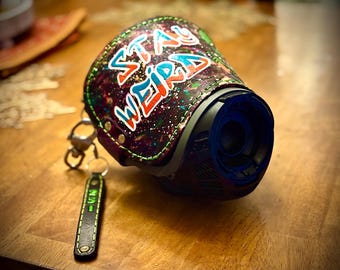 May include: A black and blue device with a leather cover featuring the words "STAY WEIRD" in colorful graffiti-style lettering. A small black leather keychain with green text hangs from the device.